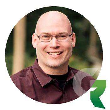 A smiling man with a shaved head and glasses wearing a dark button-down shirt.