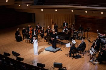 A singer and a speaker perform on a stage in front of a chamber orchestra in a concert hall.