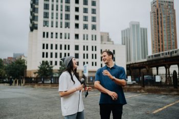 A woman holding a microphone interviews a man in a parking lot with city buildings in the background.