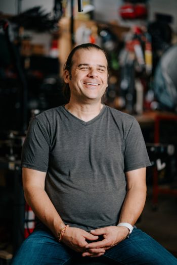 A smiling man in a grey v-neck t-shirt sitting in a workshop.