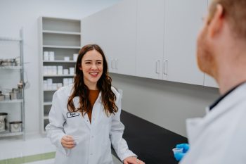 A smiling woman in a lab coat holding a small sample cup talks to a person in a lab coat in a testing facility.