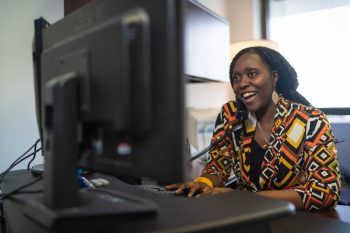 A woman smiling while talking on a telephone at her desk in front of a computer monitor.