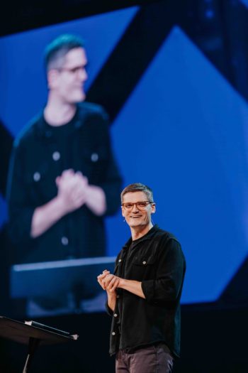 A man in glasses and a black shirt stands on a stage, with his image projected on a large screen behind him.