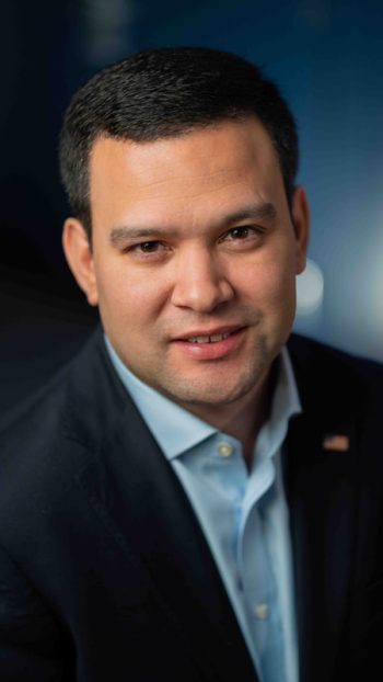 Headshot of a man with short dark hair wearing a blue button-down shirt and a dark blazer.