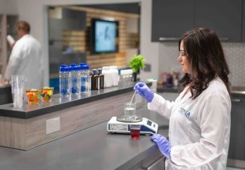A scientist in a lab coat and gloves stirs a liquid in a beaker on a digital scale.