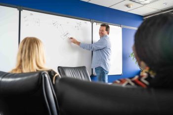 A man presents a flowchart drawn on a whiteboard to two colleagues in an office.