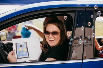 A woman in sunglasses smiles from the driver’s seat of a car while holding a flyer for a cornhole game.