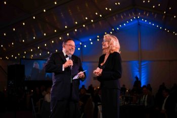 A man in a tuxedo holding a microphone and a woman in a black suit standing on a stage at an evening event.
