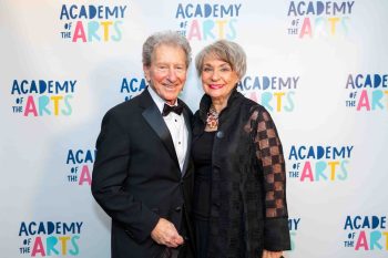 An older man in a tuxedo and a woman in a black jacket standing in front of an Academy of the Arts step and repeat wall.
