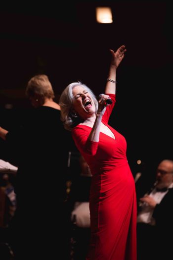 A woman in a red dress singing passionately into a microphone on stage with an orchestra behind her.