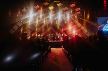 A band performs on a stage in front of a seated audience, with bright stage lights overhead.