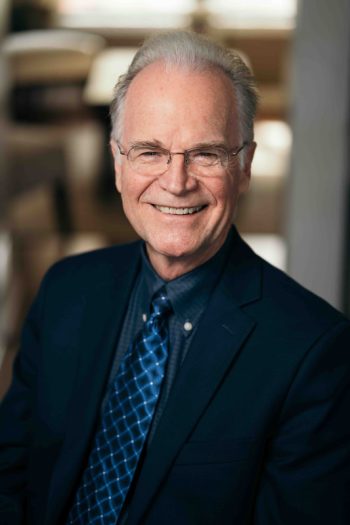 Portrait of a smiling man wearing glasses, a dark blazer, a dress shirt, and a blue patterned tie.