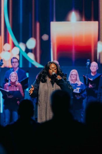 A woman singing into a microphone on stage, backed by a choir.
