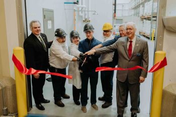 Six people wearing safety gear hold scissors together to cut a ceremonial red ribbon across a warehouse doorway.