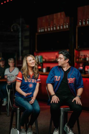 A man and a woman in Chicago Cubs jerseys sit on stools in a dimly lit bar.
