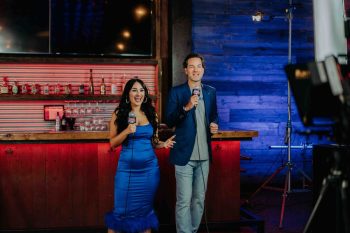 A man and a woman in a blue dress holding microphones and speaking in front of a bar.