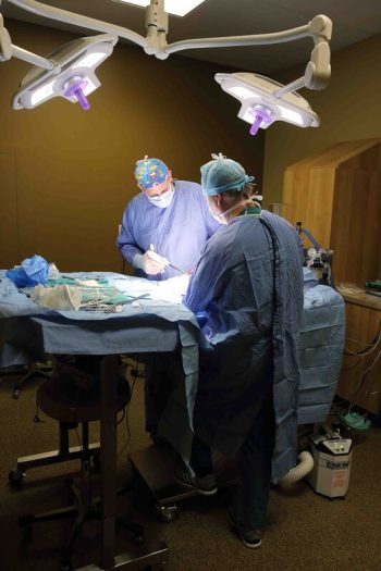 Two surgeons in scrubs performing surgery on a patient on an operating table under medical lights.