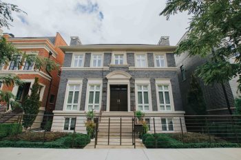 Three-story gray brick house with a dark front door, white window trim, and a metal fence in front.